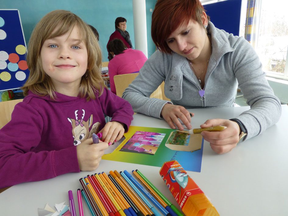 Eine Frau und ein Kind sitzen an einem Tisch, auf dem viele farbige Buntstifte liegen.