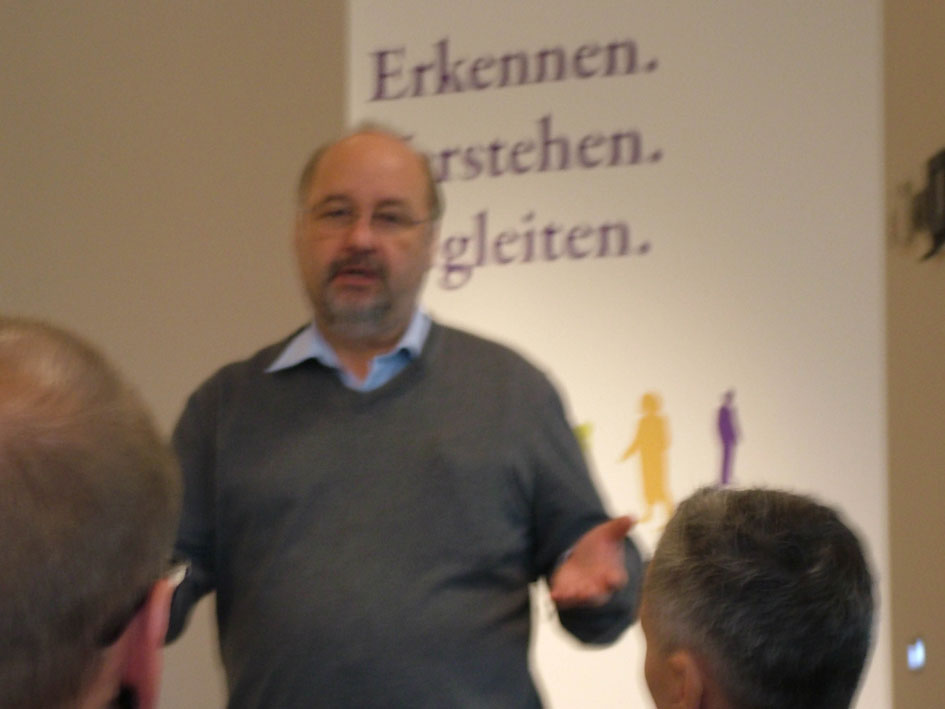 Ein Mann hält eine Rede vor einer versammelten Gruppe in einem Raum.