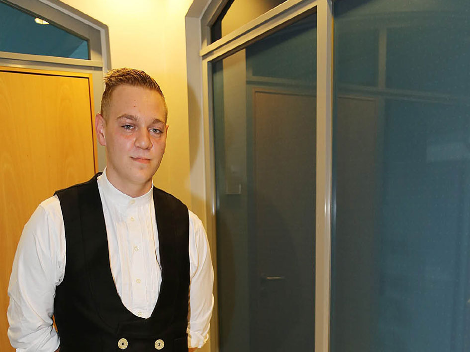 Ein Mann in einem schwarzen Weste und Krawatte steht vor einer Tür.