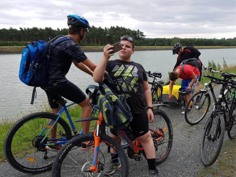 Fahrradtour am Kanal: Gruppe Jugendlicher macht Pause, einer fotografiert. Fahrräder, Rucksäcke, Freizeitaktivität, Natur, Sommer.