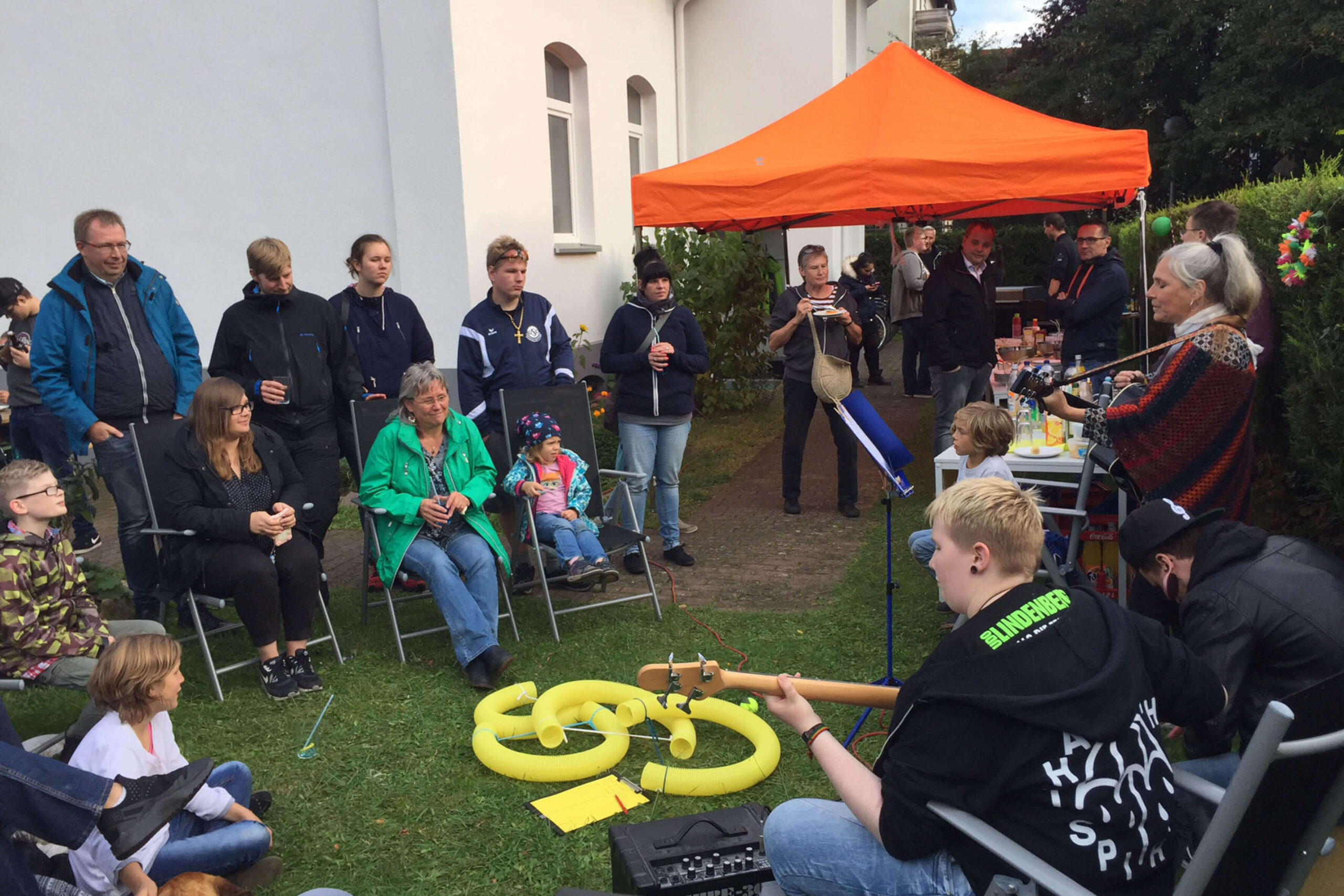 Gartenparty mit Musik: Gruppe von Menschen sitzt und steht im Garten, während Musiker Gitarre spielen und singen. Zelt im Hintergrund.