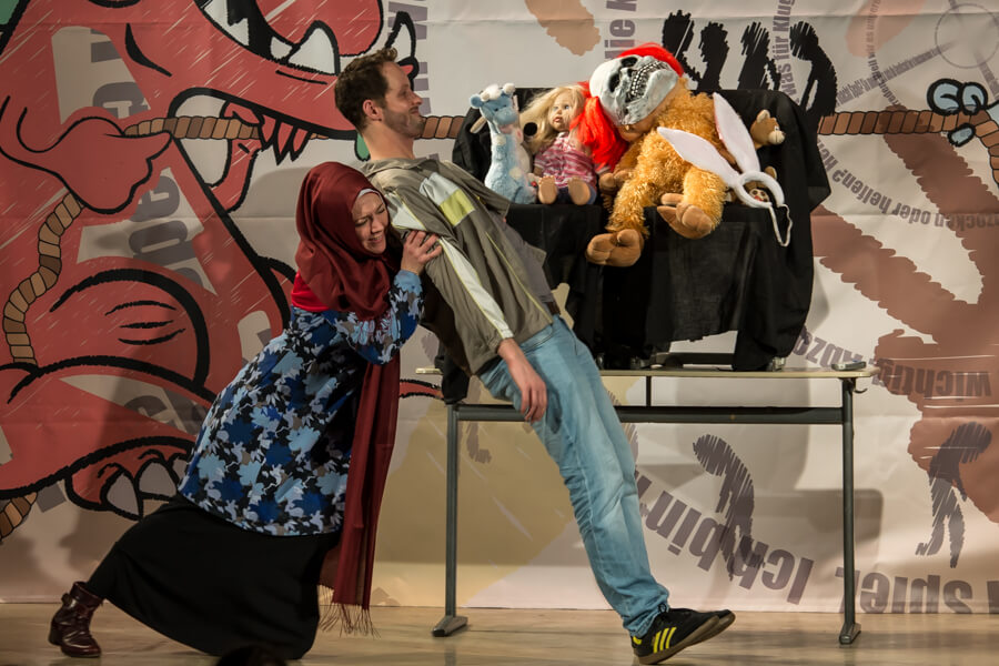 Theateraufführung: Frau mit Kopftuch und Mann ziehen an einem Seil, Puppen im Hintergrund, Bühnenbild mit Monster.
