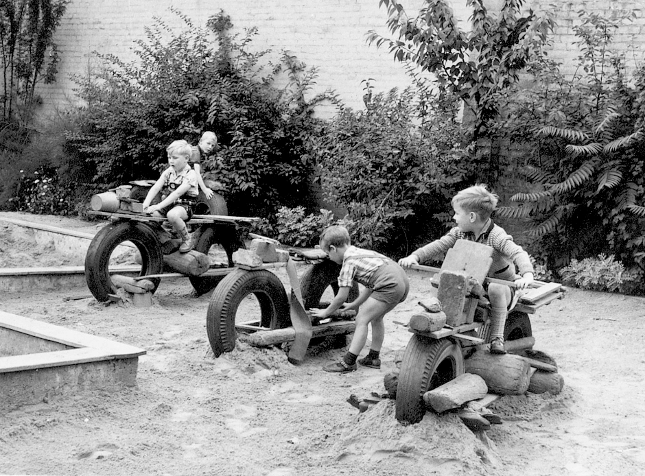 Schwarzweißfoto: Kinder spielen im Sandkasten mit selbstgebauten Motorrädern aus Reifen und Holz, Kindheit, Spielplatz.