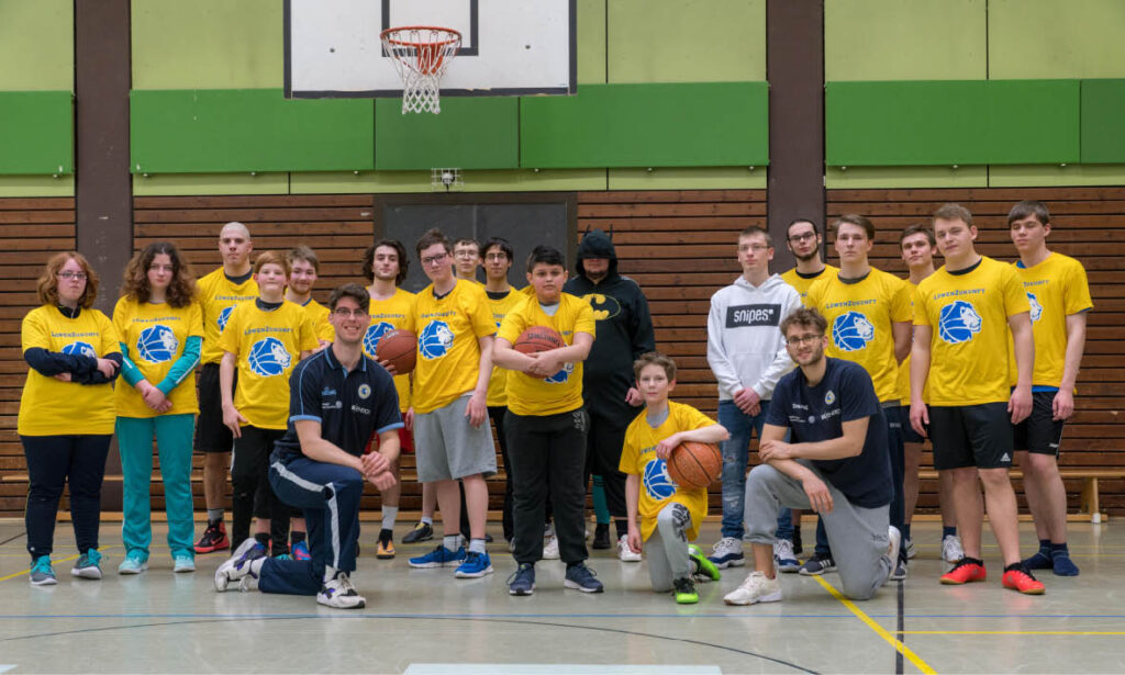 Basketballteam "Löwenpower" posiert in gelben T-Shirts mit Basketball vor Korb in Sporthalle, Trainer knien vorne.