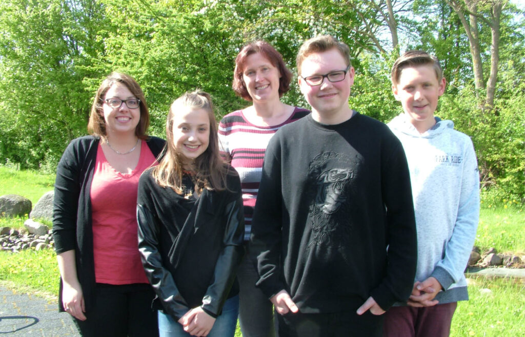 Gruppe von fünf Personen posiert im Freien vor Bäumen, Familienfoto, Generationen, lächelnde Gesichter, Freizeitaktivität.