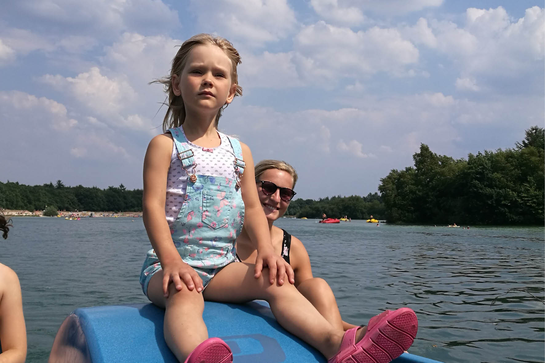 Mädchen sitzt auf Tretboot am See mit Mutter im Hintergrund. Sommerurlaub, Freizeitaktivitäten, Badesee, Wassersport.