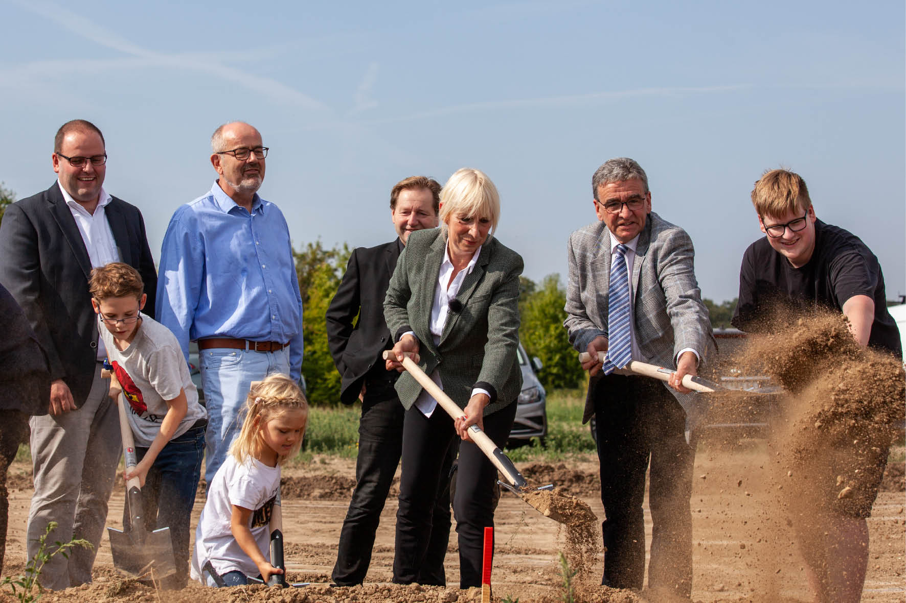 Symbolischer Spatenstich: Gruppe von Personen, darunter Politiker und Kinder, mit Spaten auf einer Baustelle für ein neues Projekt.