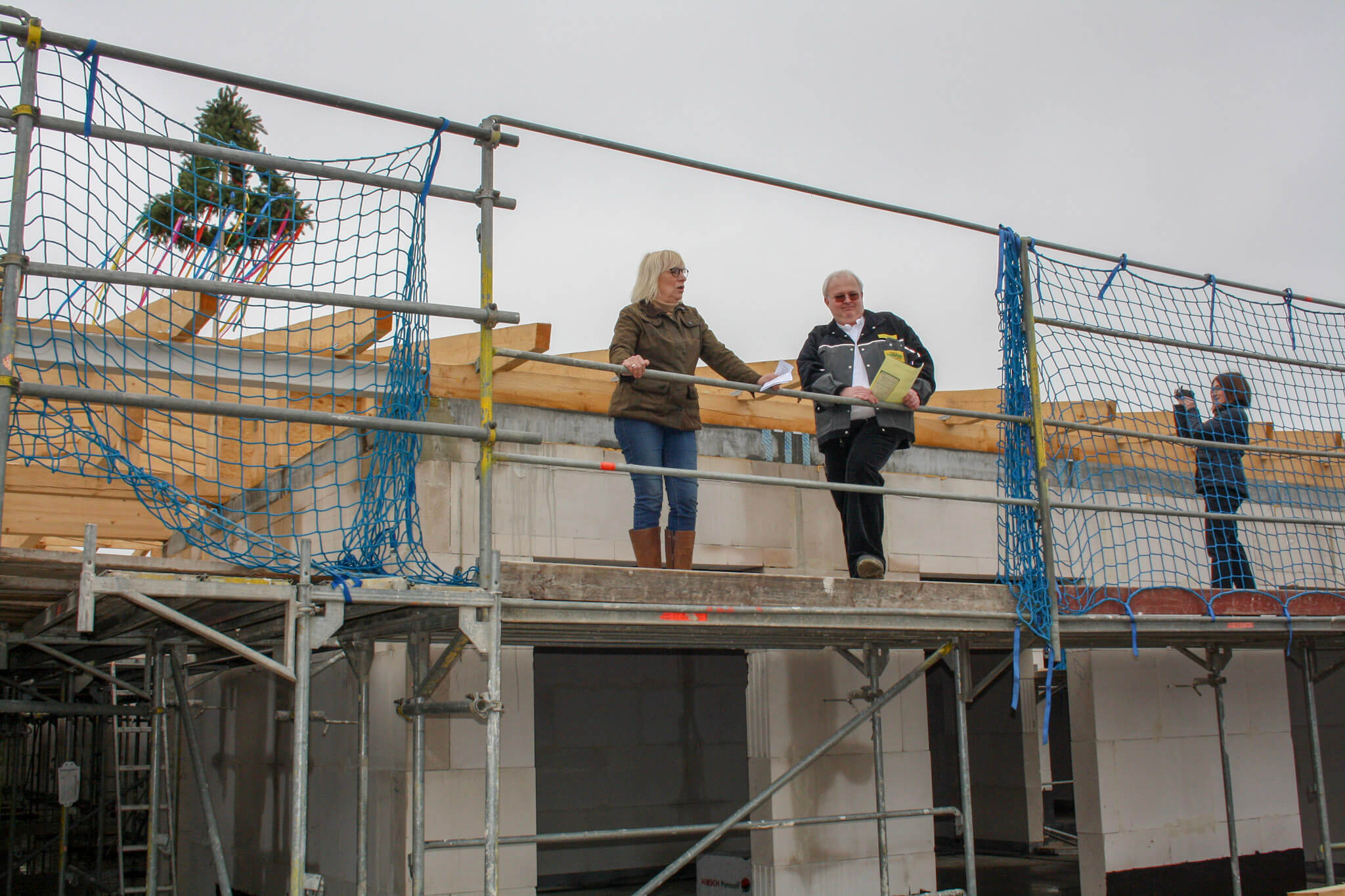 Richtfest: Bauarbeiter und Bauherren auf Gerüst am Rohbau mit Richtbaum. Neubau-Projekt, Hausbau mit Sicherheitsnetz.