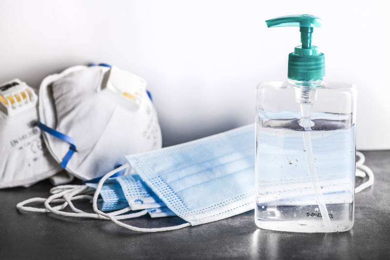 Eine Flasche mit durchsichtigem Handdesinfektionsmittel mit Pumpe steht auf einer grauen Fläche neben mehreren blauen Einweggesichtsmasken und zwei weißen N95-Masken, alles vor einem weißen Hintergrund.