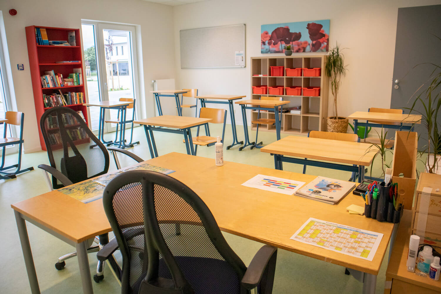 Ein helles, modernes Klassenzimmer mit auseinander stehenden Tischen und Stühlen, einem Lehrerpult mit Material, Bücherregalen, roten Ablagefächern, einem großen Fenster und einer Pflanze in der Ecke.