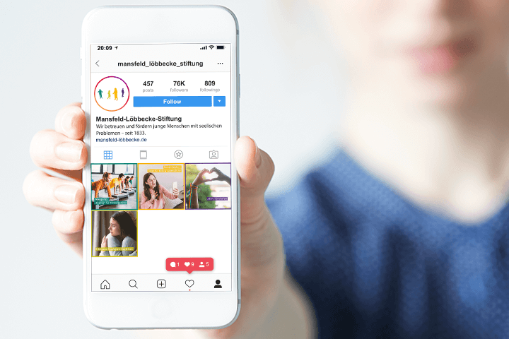 Eine Person hält ein Smartphone mit dem Instagram-Profil der mansfeld_löbbecke_stiftung in der Hand, das das Profilfoto, die Biografie und mehrere bunte Beiträge zeigt. Der Bildschirm ist scharf, während der Hintergrund unscharf ist.