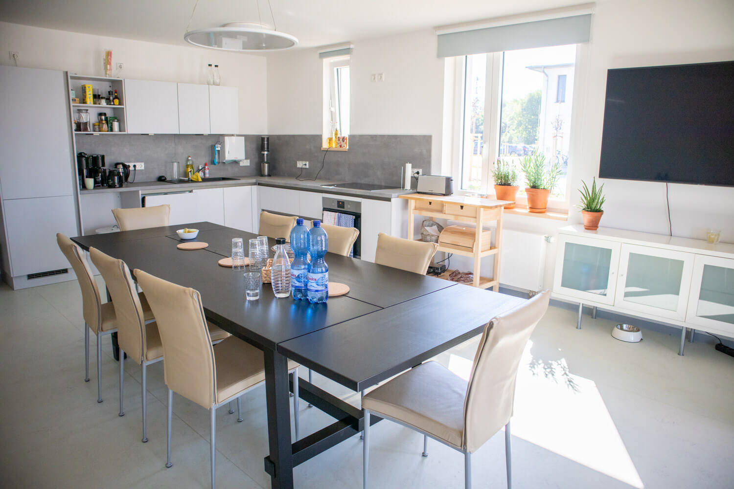 Helle, moderne Küche und Essbereich mit einem langen Esstisch, beigen Stühlen, weißen Schränken, Topfpflanzen am Fenster und einem großen Fernseher an der Wand. Flaschen und Gläser stehen auf dem Tisch. Das Sonnenlicht füllt den Raum.