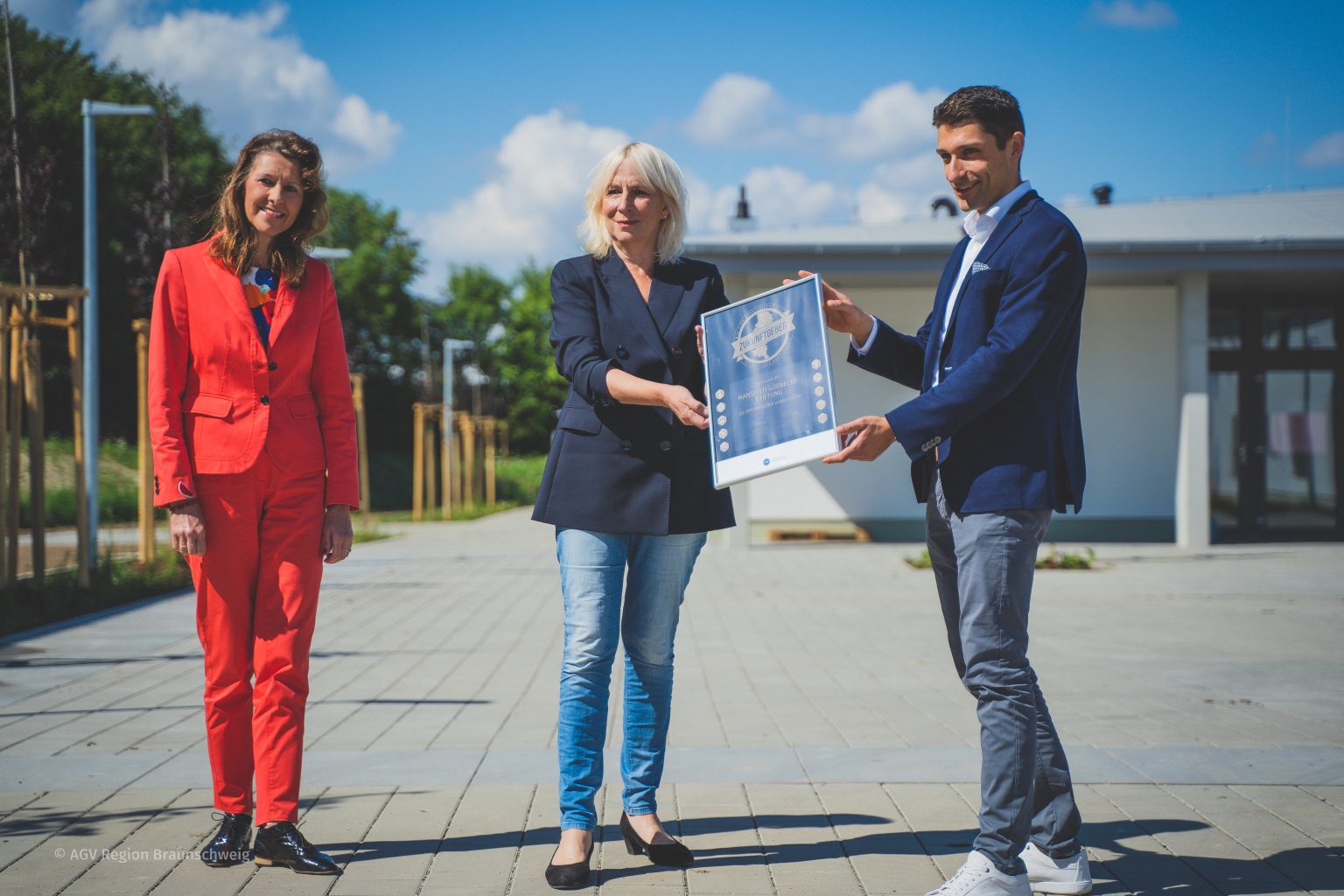 Drei Personen stehen an einem sonnigen Tag draußen; zwei von ihnen, eine Frau in einem schwarzen Blazer und ein Mann in einer blauen Jacke, halten eine gerahmte Urkunde, während eine andere Frau in einem roten Anzug lächelnd daneben steht.