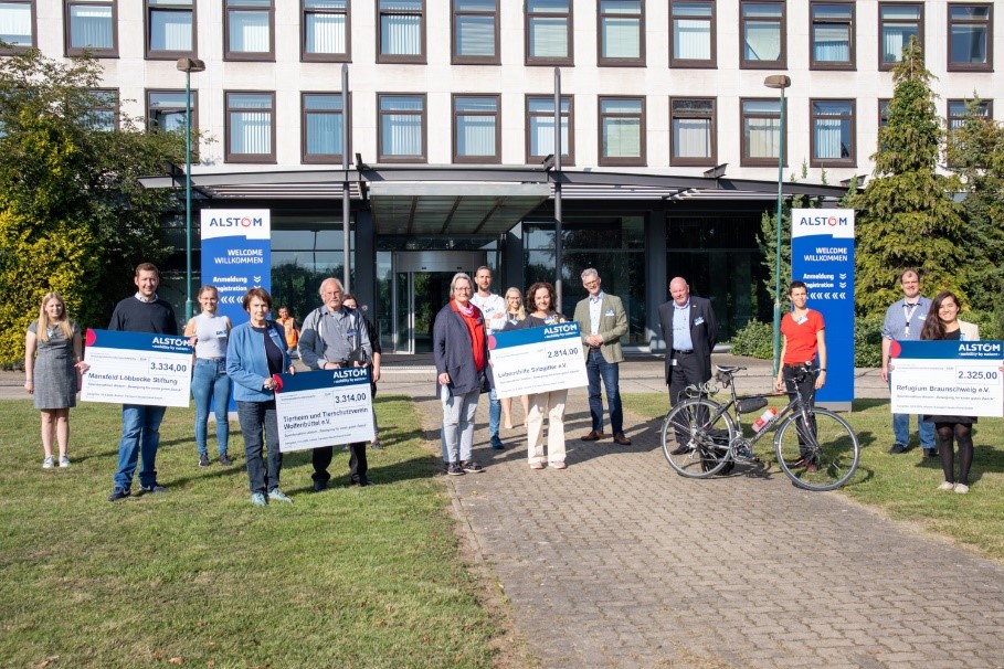 Eine Gruppe von Menschen steht vor einem Gebäude mit großen Spendenschecks in der Hand, davor zwei Fahrräder. Blaue Schilder mit dem Schriftzug ALSTOM sind in der Nähe des Eingangs zu sehen. Das Wetter ist sonnig und klar.