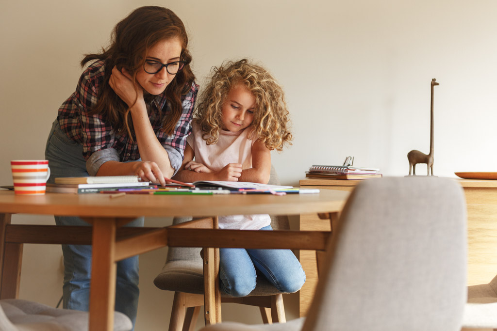 Eine Frau und ein junges Mädchen sitzen an einem Holztisch und sehen sich Papiere und Buntstifte an. Die Frau beugt sich vor, um dem Mädchen zu helfen, das sich gesetzt hat. Eine Tasse, Hefte und eine Giraffenfigur stehen auf dem Tisch.