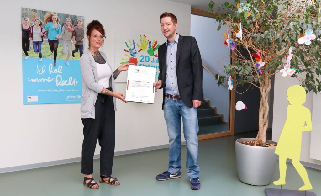 Eine Frau und ein Mann stehen in einem Haus, lächeln und halten eine Urkunde in der Hand. Im Hintergrund sind ein Plakat, ein geschmückter Baum und die gelbe Silhouette eines Kindes zu sehen.
