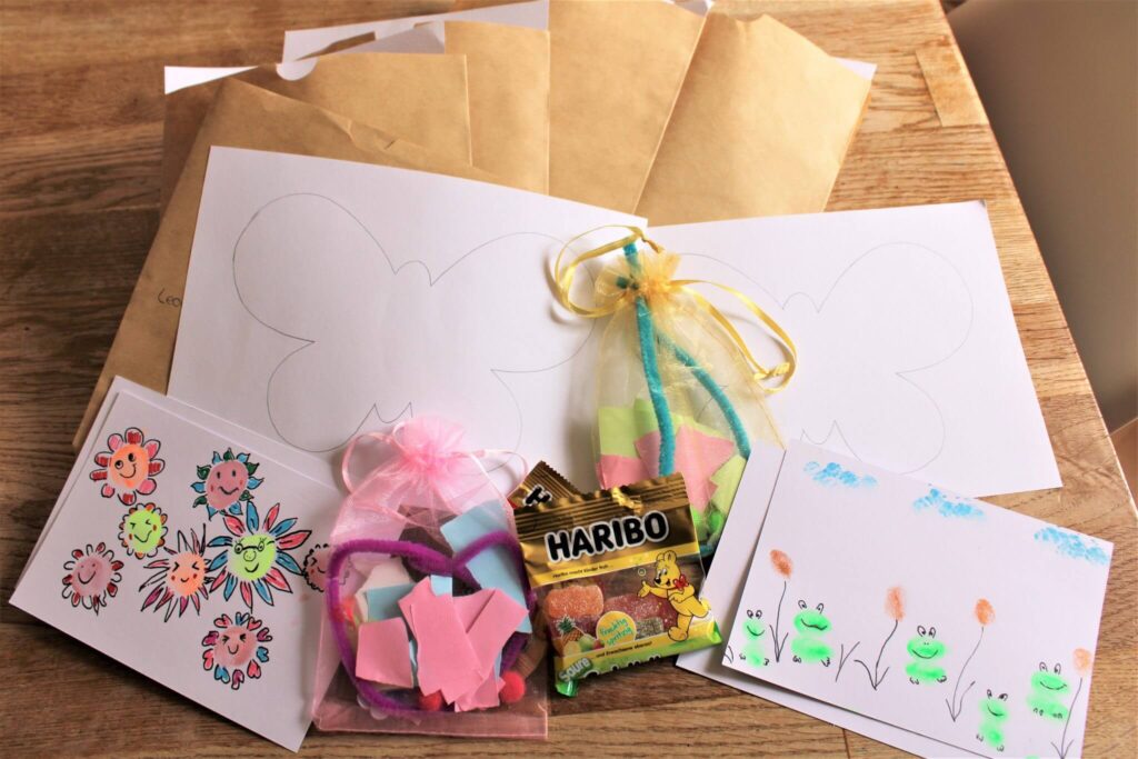 Auf einem Holztisch liegen Briefumschläge, Malblätter mit Schmetterlingen und Blumen, zwei verzierte Karten, Schaumstoffbasteleien in Netzbeuteln und eine kleine Packung Haribo-Gummibärchen.