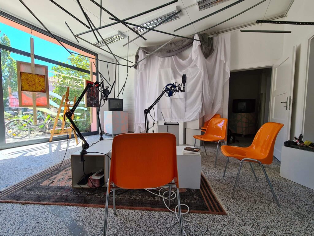 Ein modernes, hell erleuchtetes Aufnahmestudio mit orangefarbenen Stühlen, Mikrofonen auf Ständern, einer weißen Trennwand, über der Stoff drapiert ist, und großen Fenstern, durch die man Fahrräder und Grünanlagen sehen kann.