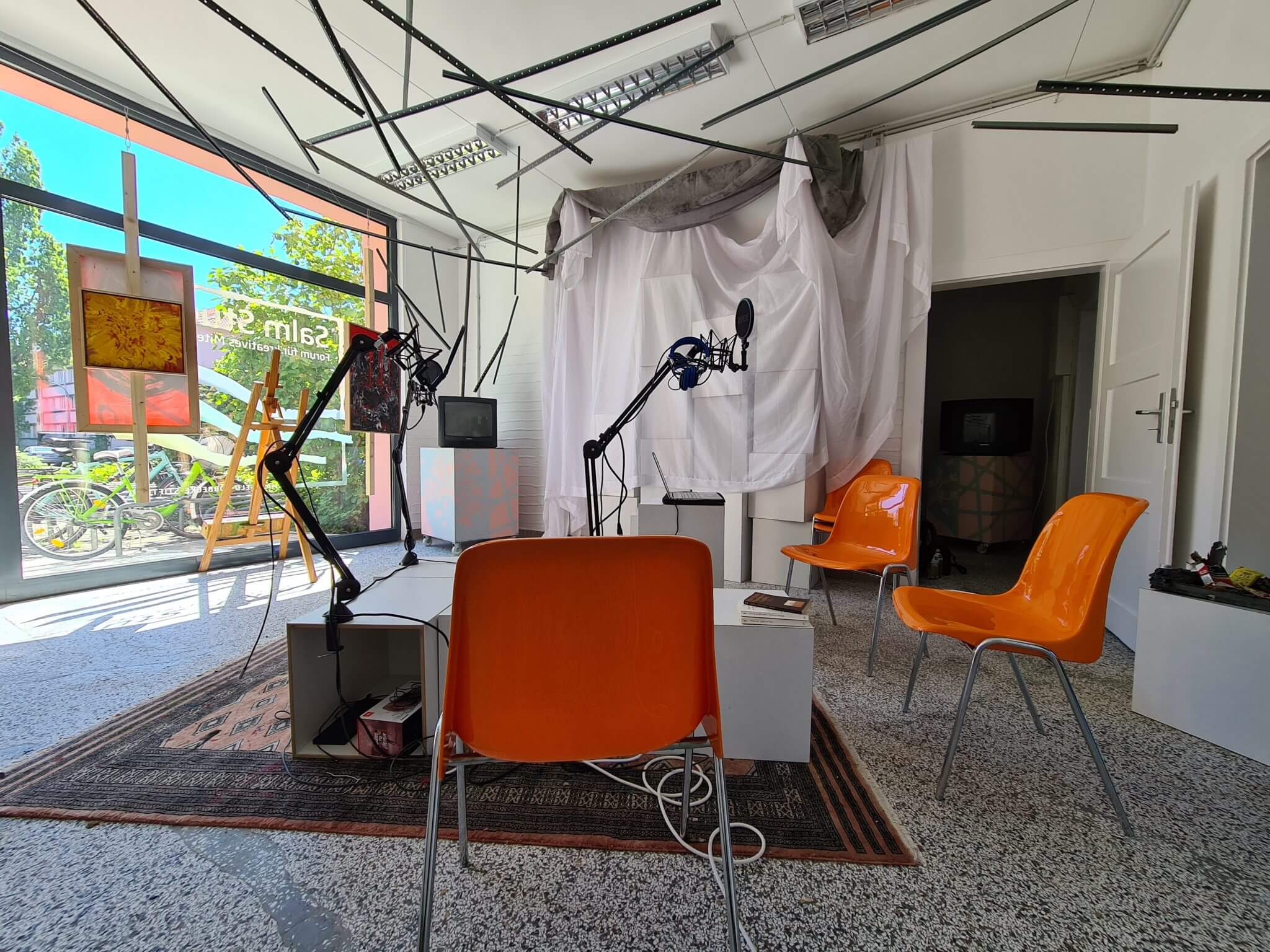 Ein modernes, hell erleuchtetes Aufnahmestudio mit orangefarbenen Stühlen, Mikrofonen auf Ständern, einer weißen Trennwand, über der Stoff drapiert ist, und großen Fenstern, durch die man Fahrräder und Grünanlagen sehen kann.