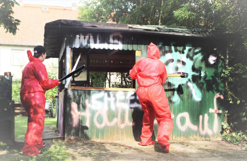 Zwei Personen in roten Schutzanzügen und schwarzen Handschuhen säubern oder überprüfen Graffiti an einem grünen Schuppen, der von Bäumen umgeben ist, und an dessen Wand gesprühte Wörter und Markierungen zu sehen sind.