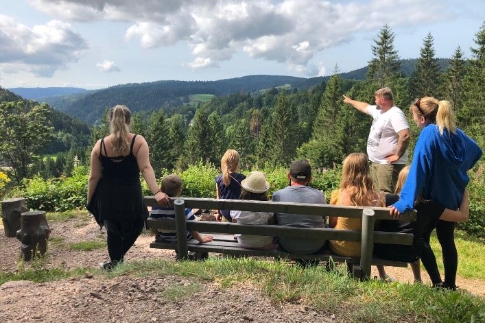 Eine Gruppe von Menschen, darunter Kinder und Erwachsene, sitzt und steht um eine Bank herum und blickt auf eine malerische, bewaldete Berglandschaft. Ein Mann steht und deutet auf die fernen Hügel unter einem teilweise bewölkten Himmel.