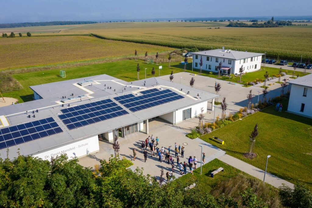 Luftaufnahme eines modernen Gebäudes mit Solarpaneelen auf dem Dach, vor dem sich eine Gruppe von Menschen versammelt hat, umgeben von grünen Rasenflächen und Feldern unter einem klaren Himmel. In der Nähe ist ein weiteres weißes Gebäude zu sehen.