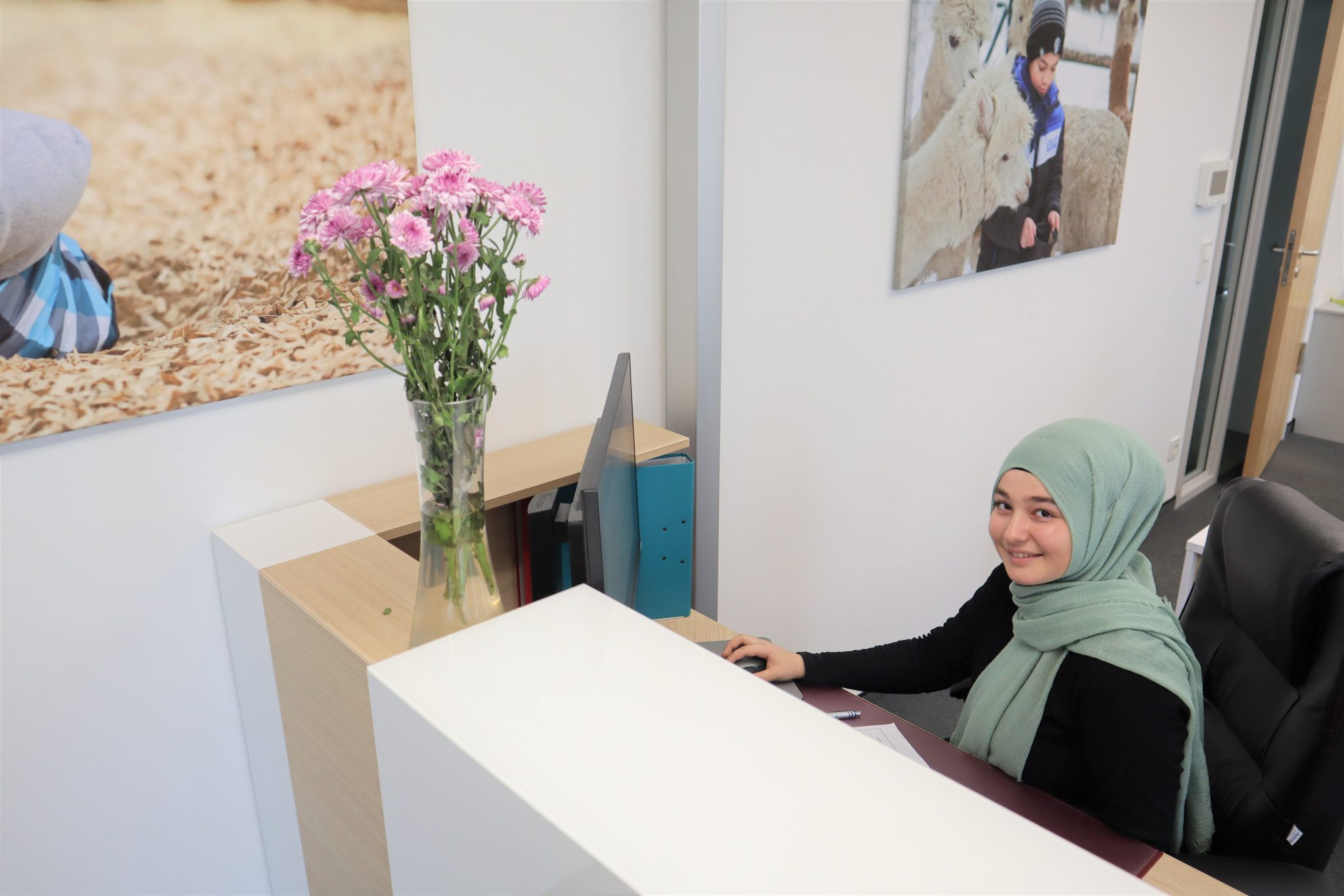 Eine Frau, die einen hellgrünen Hidschab trägt, sitzt an einem Empfangstresen und lächelt in die Kamera. Auf dem Tresen steht eine Vase mit rosafarbenen Blumen, und an der Wand hinter ihr hängen Fotos.
