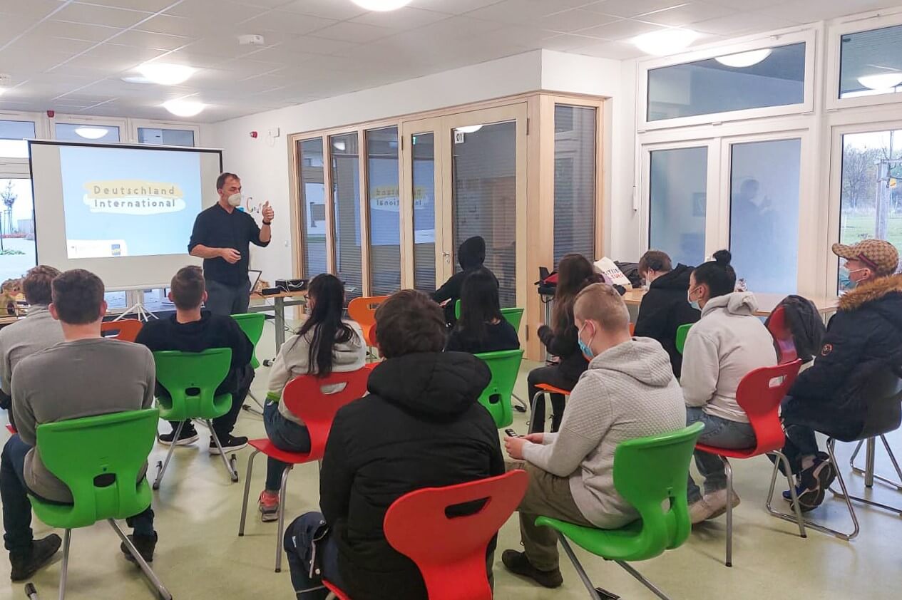 In einem hellen, modernen Klassenzimmer sitzt eine Gruppe von Menschen auf bunten Stühlen vor einem Sprecher, der neben einer projizierten Präsentation steht, auf der Deutschland International zu lesen ist.