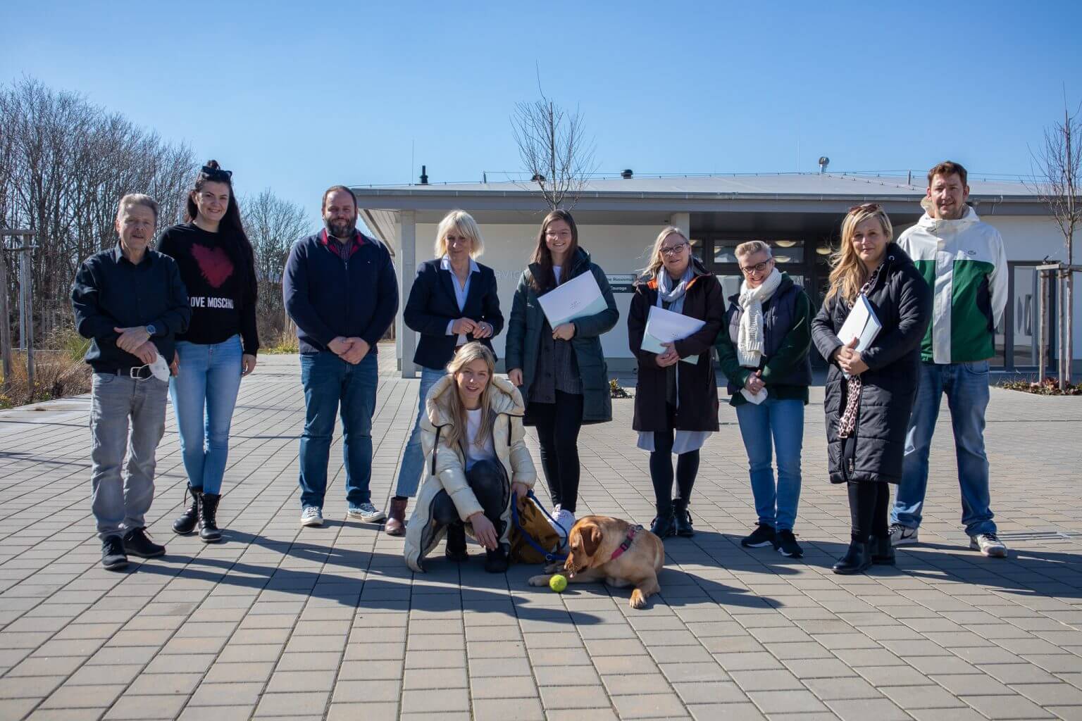Gruppe von Menschen mit Hund und Tennisball vor Gebäude. Teamfoto, Außentermin, sonniger Tag.