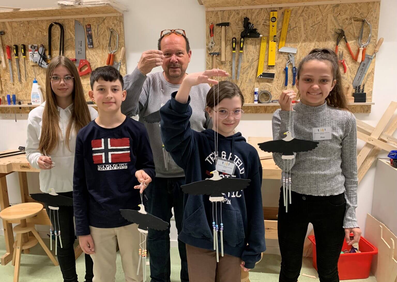 Kinder basteln Windspiele mit Adler-Motiv im Werkraum, betreut von einem Erwachsenen. Handwerksprojekt für Kinder.