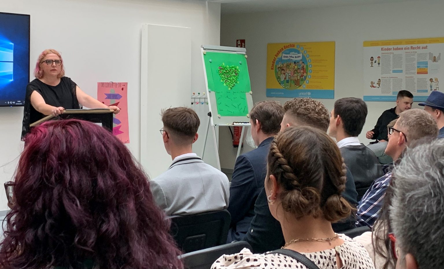 Frau hält Vortrag vor Publikum, Flipchart mit Herz, Kinderrechte Plakate im Hintergrund, Seminarraum, Bildung.