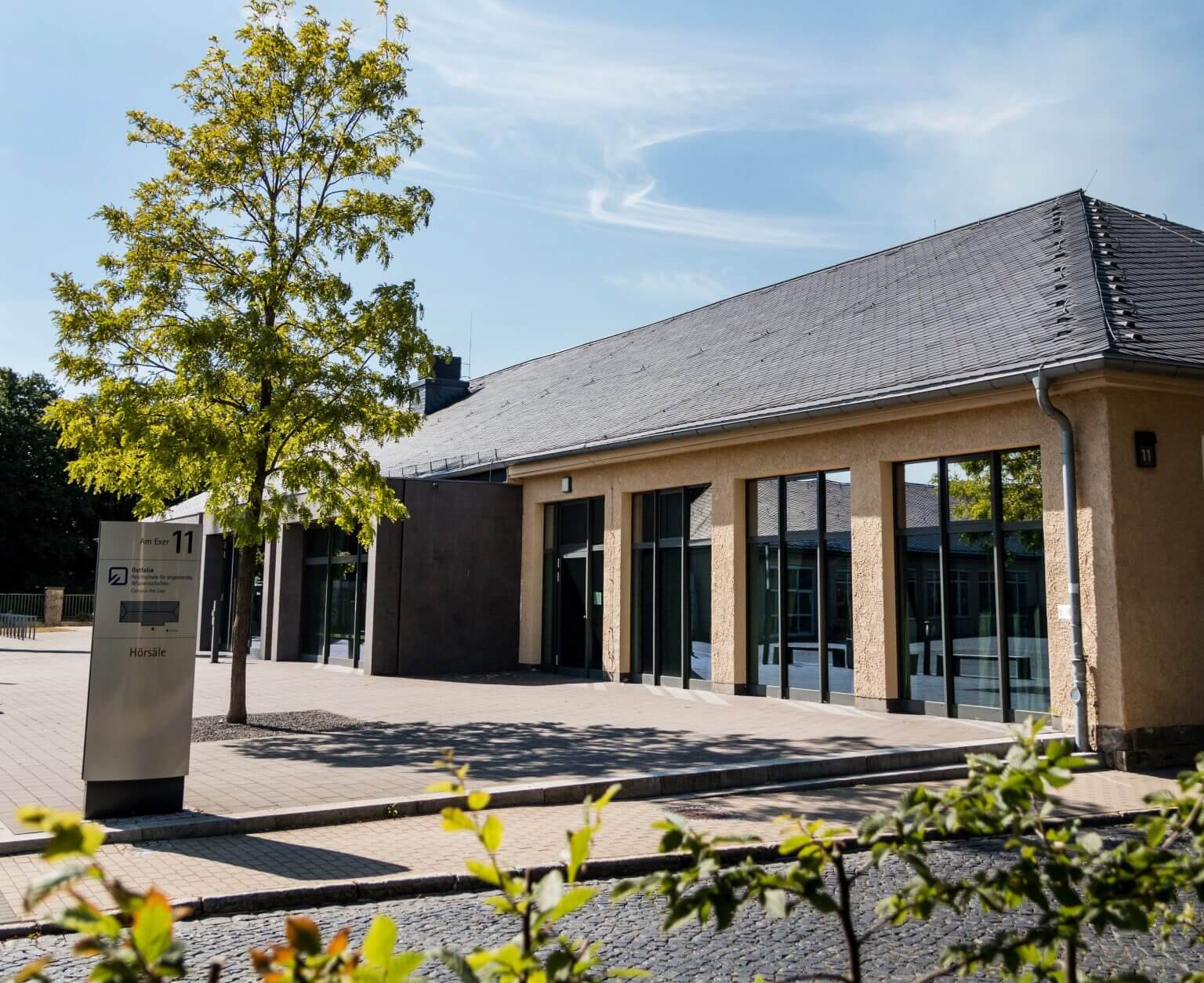 Hörsaalgebäude Universität, Campus mit Baum und Pflastersteinen. Architektur mit großen Fensterfronten und Hinweisschild.