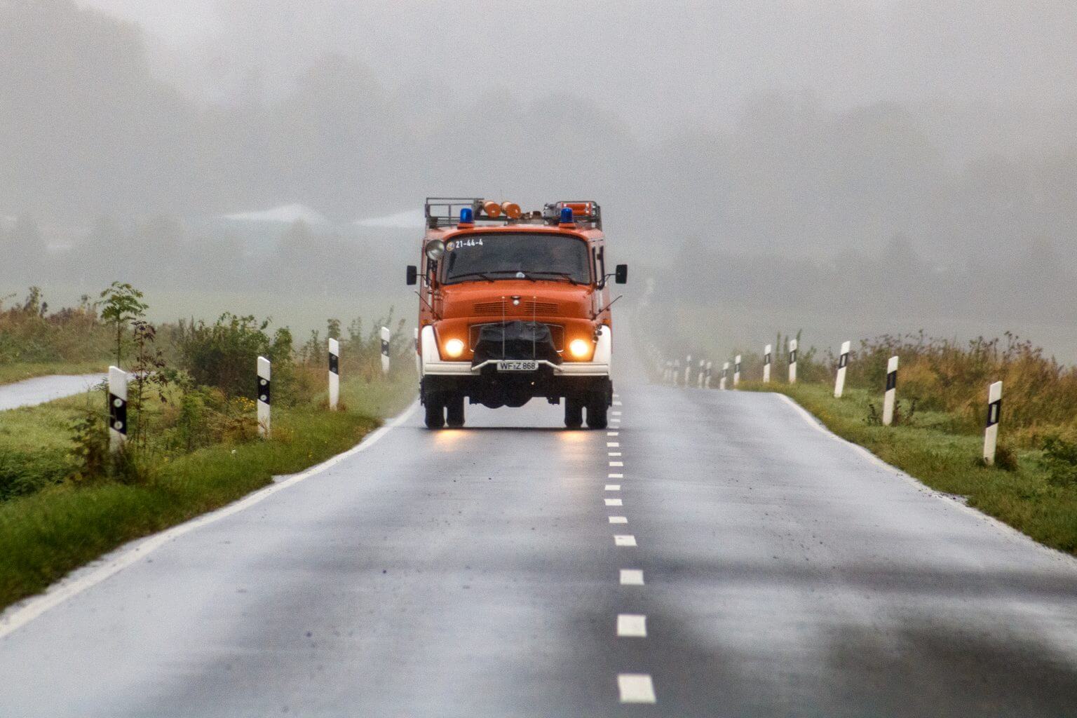 Orangerotes Feuerwehrauto fährt auf regennasser Landstraße, Blaulicht und Scheinwerfer an. Einsatzfahrzeug der Feuerwehr.