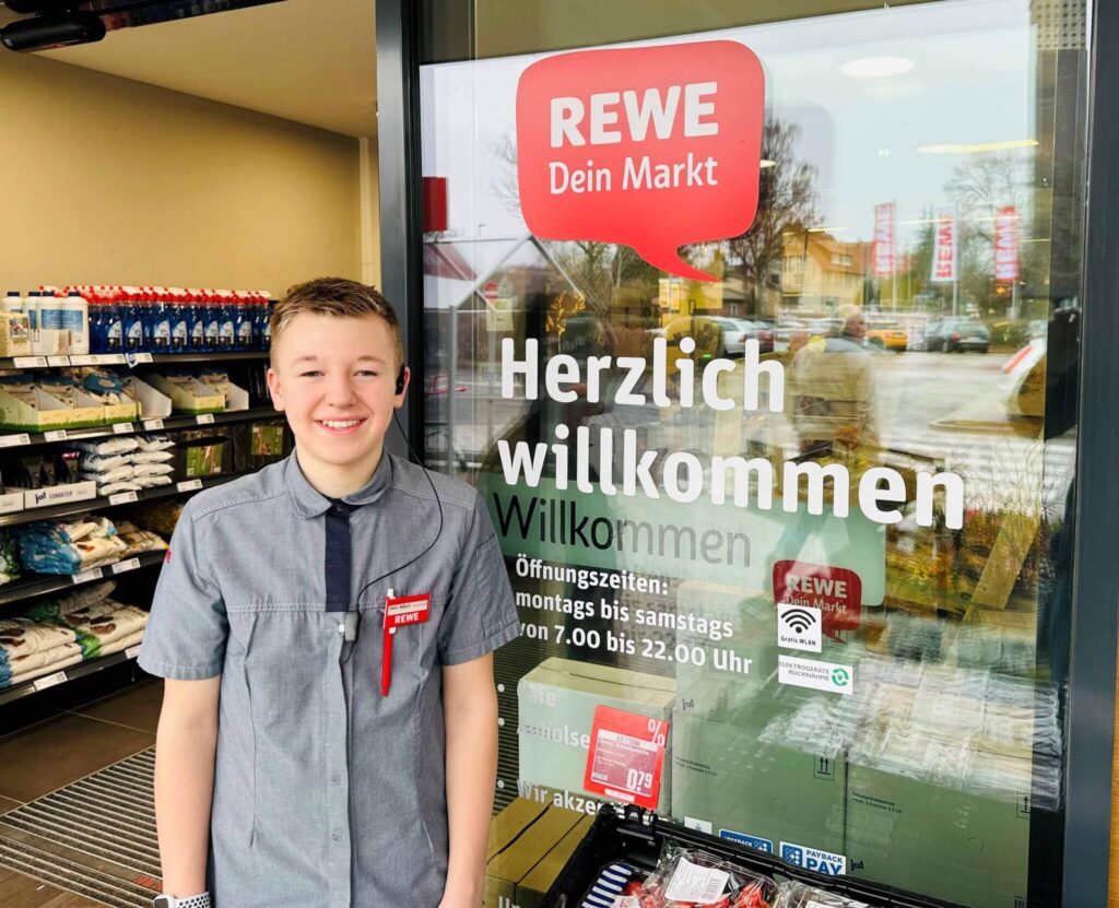 Eine lächelnde junge Person in einer REWE-Uniform steht vor dem Eingang eines REWE-Supermarktes neben einer Glastür mit der Aufschrift "Herzlich willkommen" und "Öffnungszeiten" in deutscher Sprache. Im Inneren des Ladens sind Regale und Produkte zu sehen.