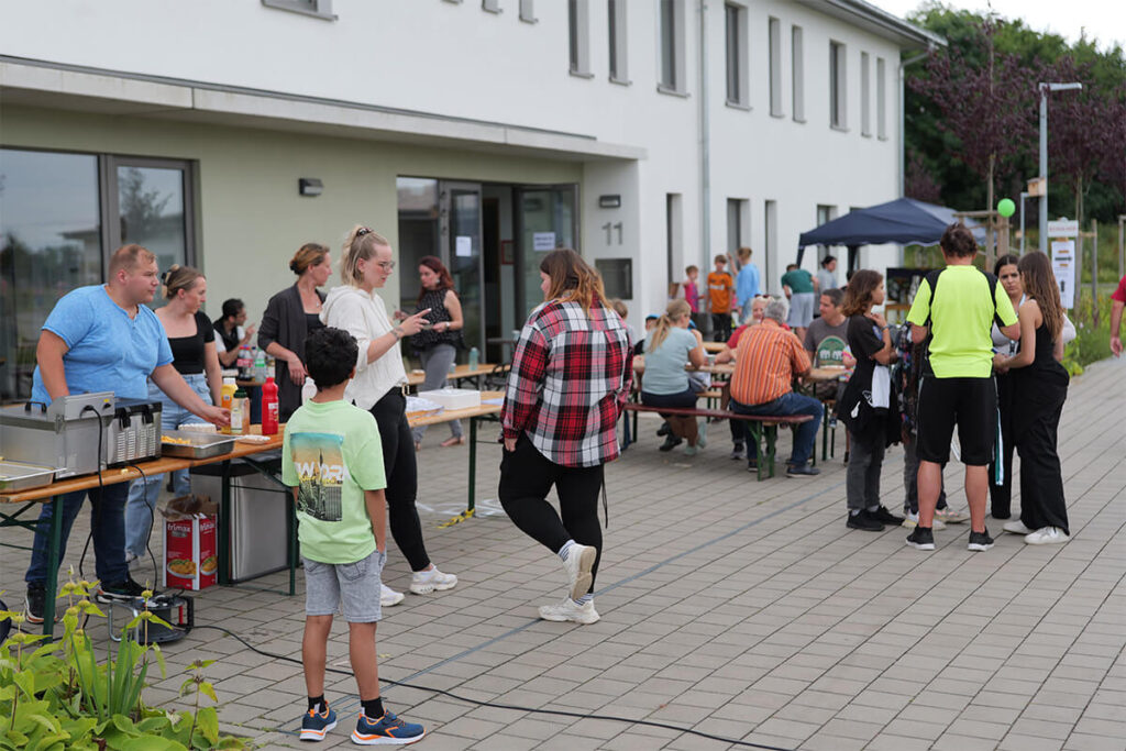 Menschen stehen um Tische mit Essen und Getränken, während sie sich unterhalten und die Atmosphäre genießen.