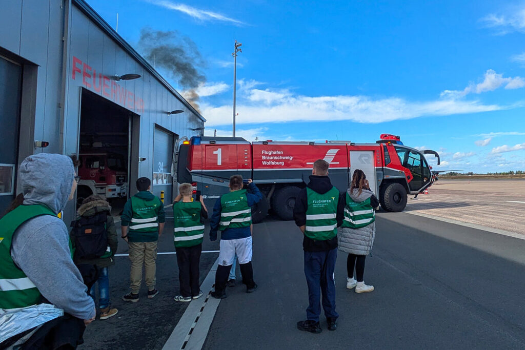 Eine Gruppe von Menschen steht lächelnd vor einem Feuerwehrauto, das im Hintergrund sichtbar ist.