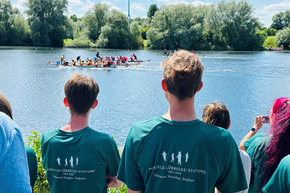 Drachenbootrennen auf dem See beobachtet von Zuschauern mit Misfield-Löbbecke-Stiftung T-Shirts. Sommerliche Outdoor-Aktivität.