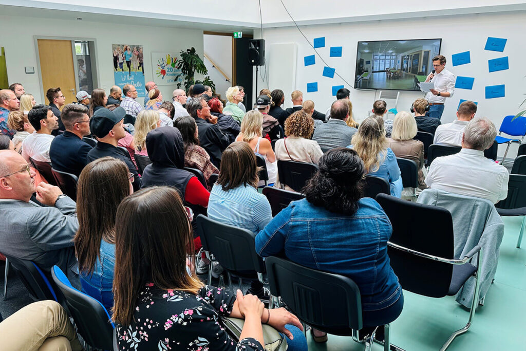 Eine große Gruppe von Menschen in einem Raum, die einer Präsentation aufmerksam zuschaut.