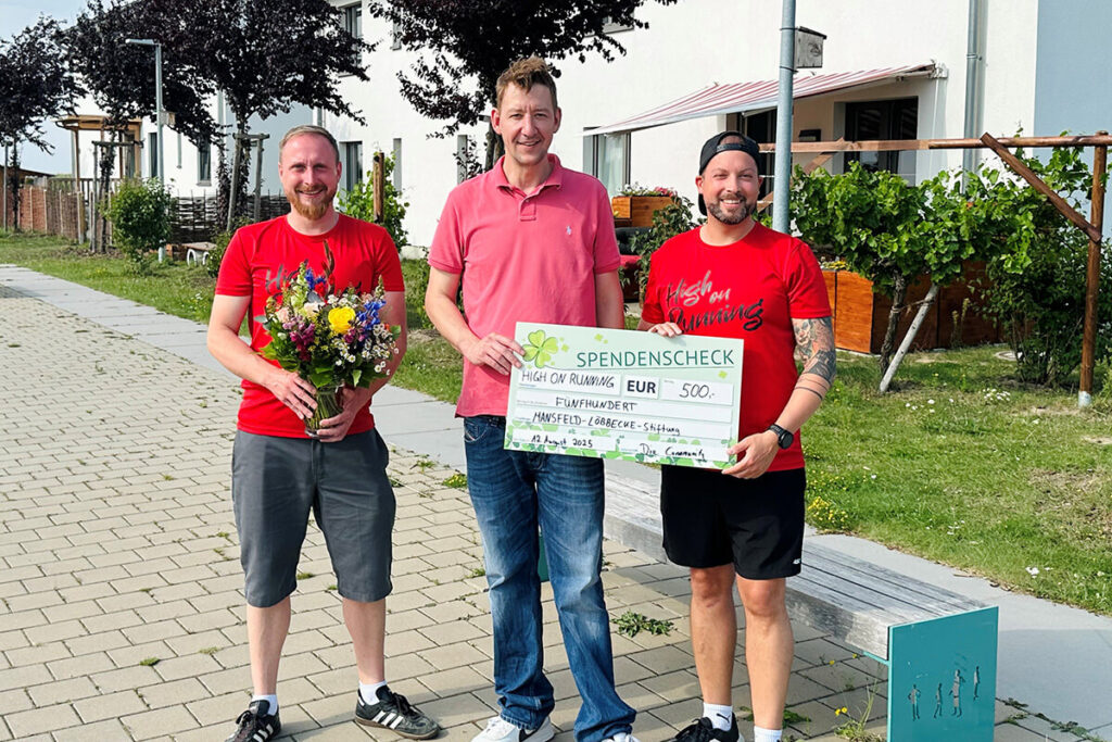 Drei Männer in roten Hemden halten einen Scheck und Blumen in den Händen.