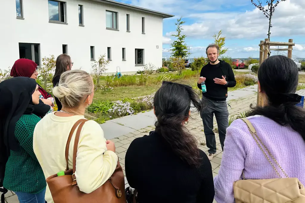 Ein Mann spricht mit einer Gruppe von Frauen auf einem gepflasterten Weg neben einem weißen Gebäude. Die Szene spielt im Freien, bei strahlend blauem Himmel und ruhiger Atmosphäre.