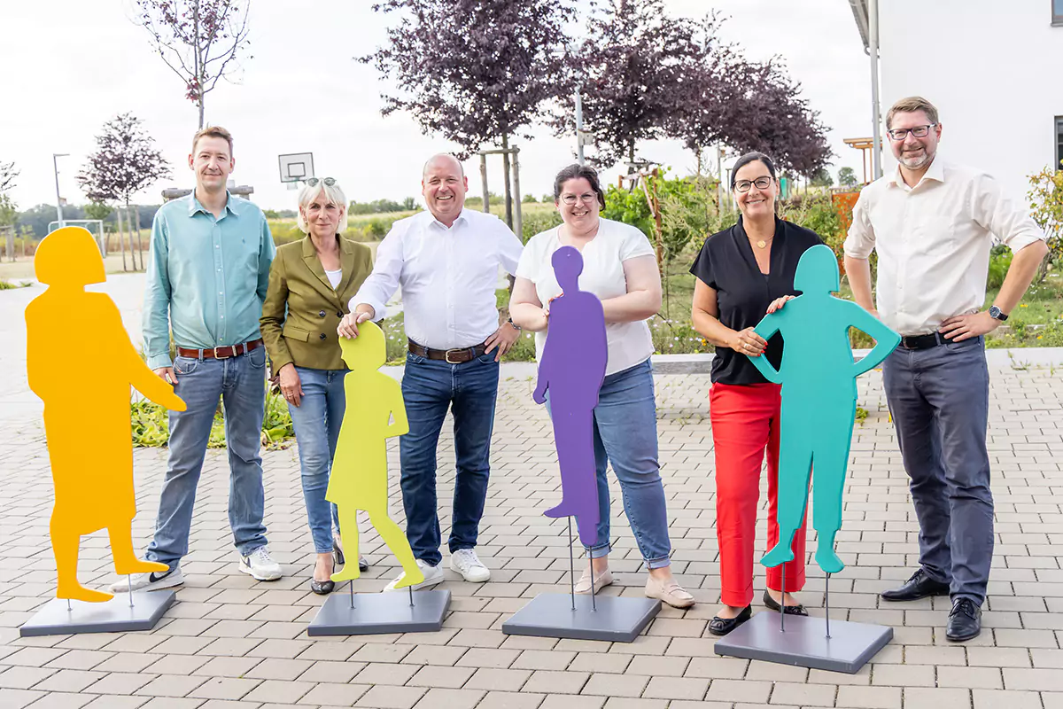 Eine Gruppe von sechs Personen steht im Freien und lächelt neben bunten Silhouettenausschnitten von Menschen.