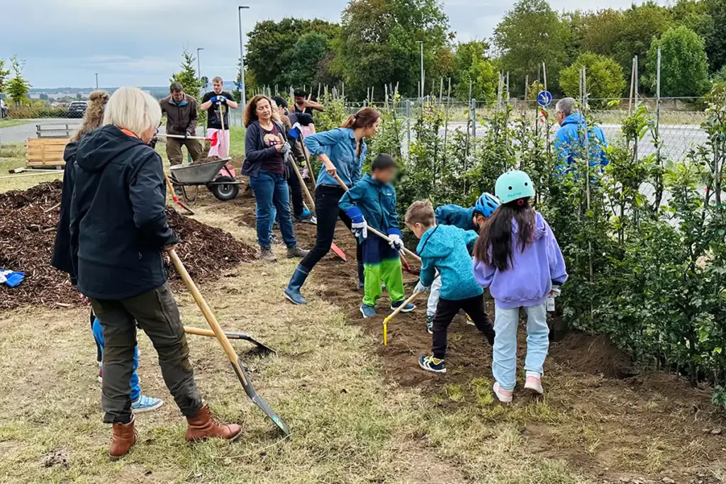 Eine Gruppe von Erwachsenen und Kindern in Freizeitkleidung und mit Helmen gräbt Erde um und pflanzt gemeinsam neben einer Reihe junger Bäume. Die Szene ist lebhaft und von Zusammenarbeit geprägt.